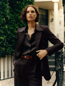 Woman in a dark suit standing outdoors with greenery and a building in the background