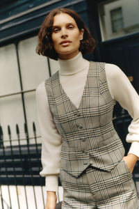 Woman wearing a gray plaid vest and pants with a white turtleneck sweater outdoors.